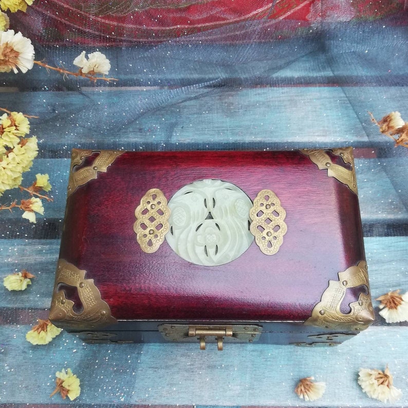 May include: A rectangular wooden box with a deep red finish, adorned with brass corner accents and a central jade carving. The box features a decorative clasp and two brass embellishments on either side of the jade carving.