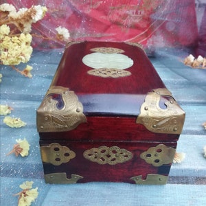 May include: A rectangular, dark red wooden box with brass corner accents and a central jade-colored oval inlay. The box has a glossy finish and ornate brass hardware. The design suggests a vintage or antique style.