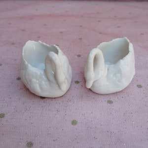 May include: Two white porcelain swan-shaped trinket dishes. The swans are facing each other with their necks curved.