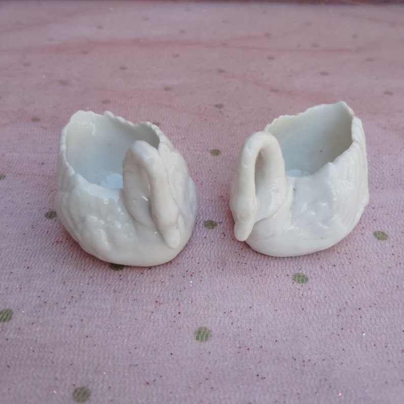 May include: Two white porcelain swan-shaped trinket dishes. The swans are facing each other with their necks curved.