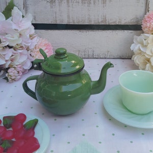 Pode incluir: Uma chaleira de esmalte verde com uma borda preta sobre um fundo branco. A chaleira está sobre uma toalha de mesa branca com bolinhas verdes.