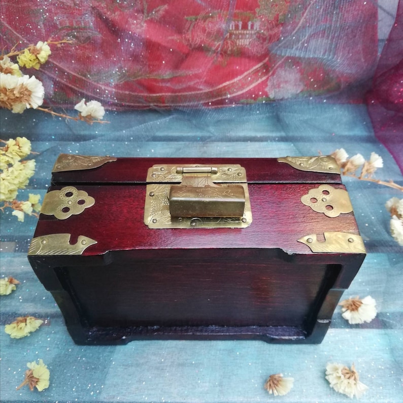 May include: A dark red wooden box with brass hardware, including corner brackets and a rectangular clasp. The box has a rectangular shape and a hinged lid. The brass hardware adds a decorative touch to the box.