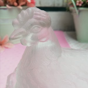 May include: A frosted glass chicken figurine with a detailed head and feathers. The chicken is sitting on a pink surface.