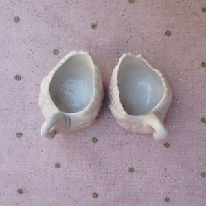 May include: Two white ceramic cups shaped like swans with their heads tilted back. The cups are empty and have a smooth, glossy finish.