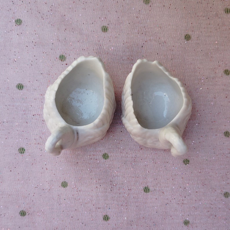 May include: Two white ceramic cups shaped like swans with their heads tilted back. The cups are empty and have a smooth, glossy finish.