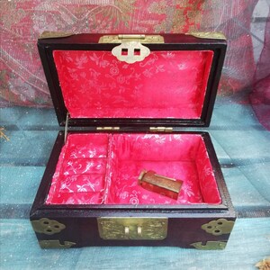 May include: Open wooden jewelry box with a dark brown exterior and brass hardware. The interior is lined with red fabric featuring a floral pattern. A small, rectangular object rests inside the box.