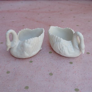 May include: Two white ceramic swan-shaped dishes. The dishes are detailed with raised feathers and have a smooth, glossy finish.