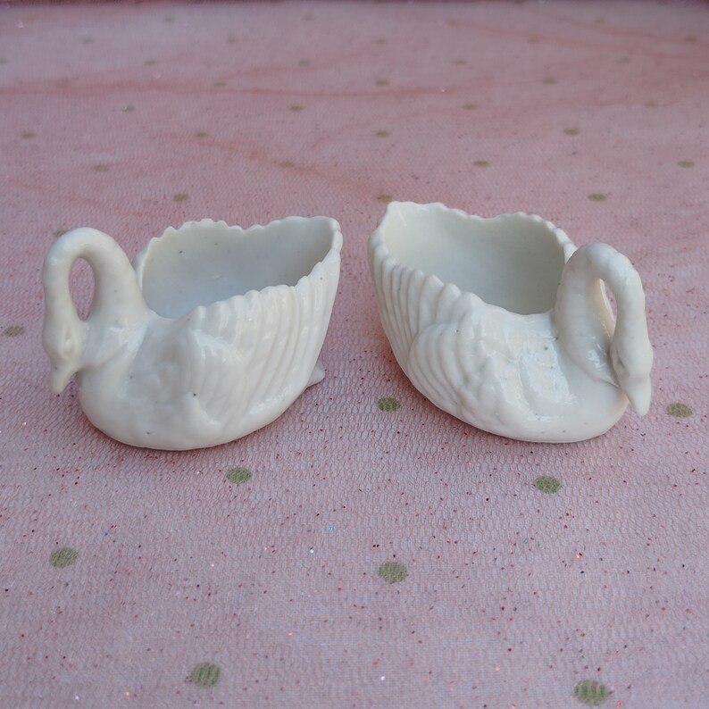 May include: Two white ceramic swan-shaped dishes. The dishes are detailed with raised feathers and have a smooth, glossy finish.