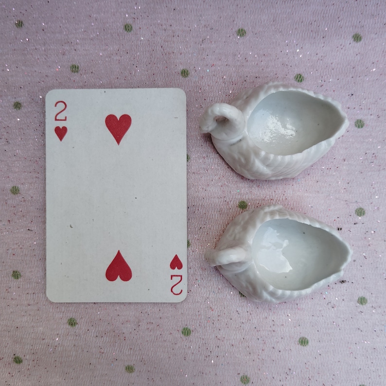 May include: Two white porcelain swan-shaped dishes with a small indentation in the centre. The dishes are on a pink background with white polka dots. A playing card with the two of hearts is also in the image.