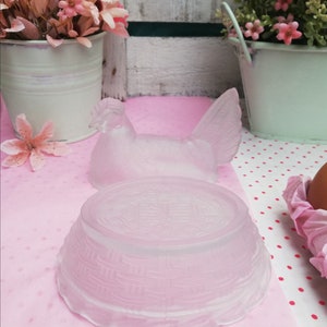 May include: A frosted glass chicken-shaped egg holder with a woven basket base. The holder is on a pink and white polka dot surface.