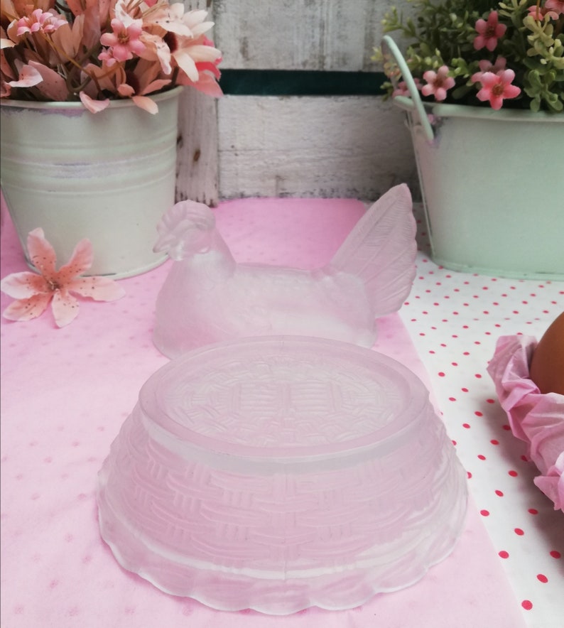 May include: A frosted glass chicken-shaped egg holder with a woven basket base. The holder is on a pink and white polka dot surface.