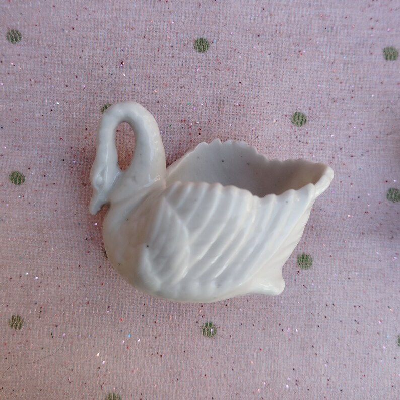 May include: A white porcelain swan planter with a detailed, textured design. The swan's neck is curved and the planter has a small opening for planting.