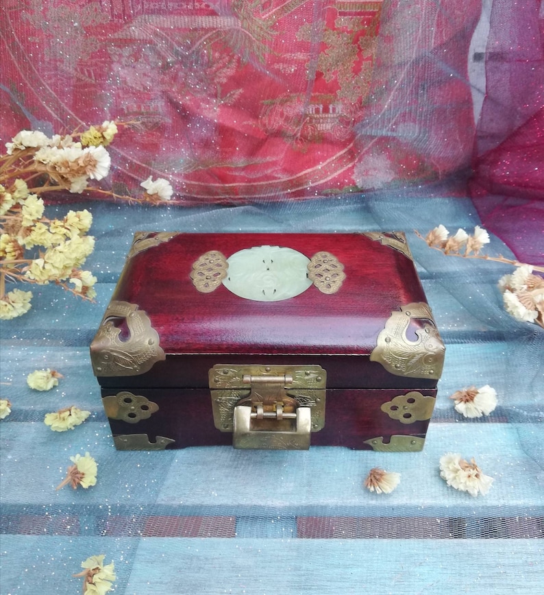 May include: A rectangular, dark red wooden box with brass corner accents and a central clasp. The lid features a light green oval jade piece and decorative brass hardware. The box is likely for jewelry or small keepsakes.