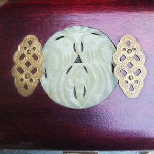 May include: Close-up of a wooden box with a carved jade centerpiece and brass accents. The jade carving features a bird design. The box has a dark red finish and decorative brass hardware.