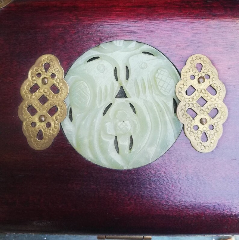 May include: Close-up of a wooden box with a carved jade centerpiece and brass accents. The jade carving features a bird design. The box has a dark red finish and decorative brass hardware.