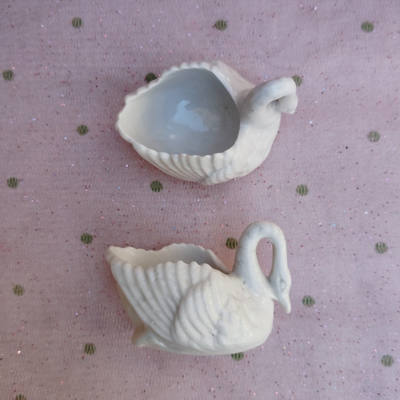 May include: Two white porcelain swan-shaped trinket dishes. The swans have detailed wings and heads.