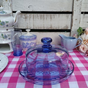 Puede incluir: Una campana de queso de cristal azul cobalto con un plato a juego, sobre un mantel a cuadros rosa y blanco. Se ven otros artículos de vajilla vintage azules y blancos, como una tetera y una jarra de crema.