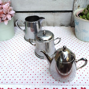 May include: Three silver metal serving pieces on a white tablecloth with red polka dots. A creamer, a sugar bowl with a lid, and a teapot.