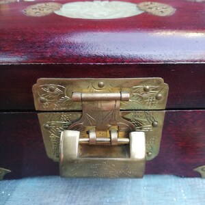 May include: A close-up of a wooden box with a deep red finish. The box features a decorative brass clasp and hinges with intricate floral designs. A light-colored, oval-shaped inlay is visible on the lid.