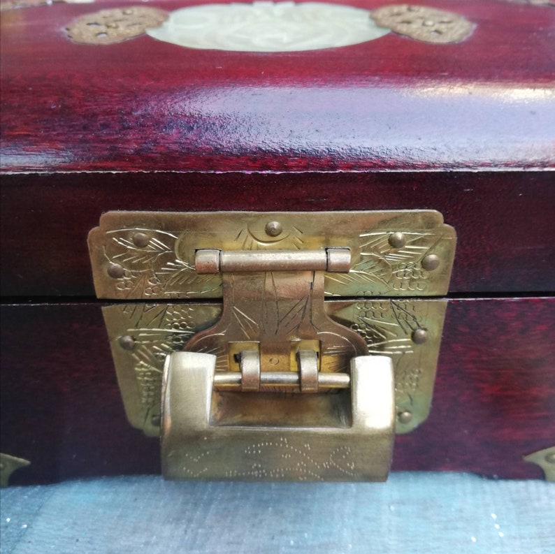 May include: A close-up of a wooden box with a deep red finish. The box features a decorative brass clasp and hinges with intricate floral designs. A light-colored, oval-shaped inlay is visible on the lid.