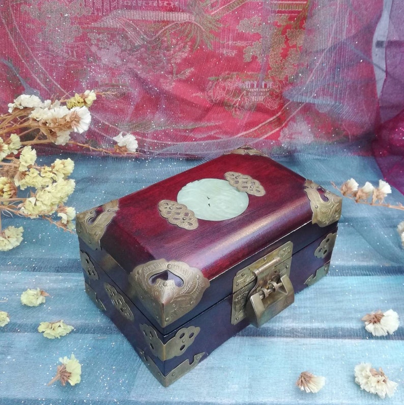 May include: A rectangular wooden box with a rich, dark red finish. The box features ornate brass corner protectors and a central jade-colored stone inlay. The lid is secured with a brass clasp, and the overall design suggests a vintage or antique aesthetic.