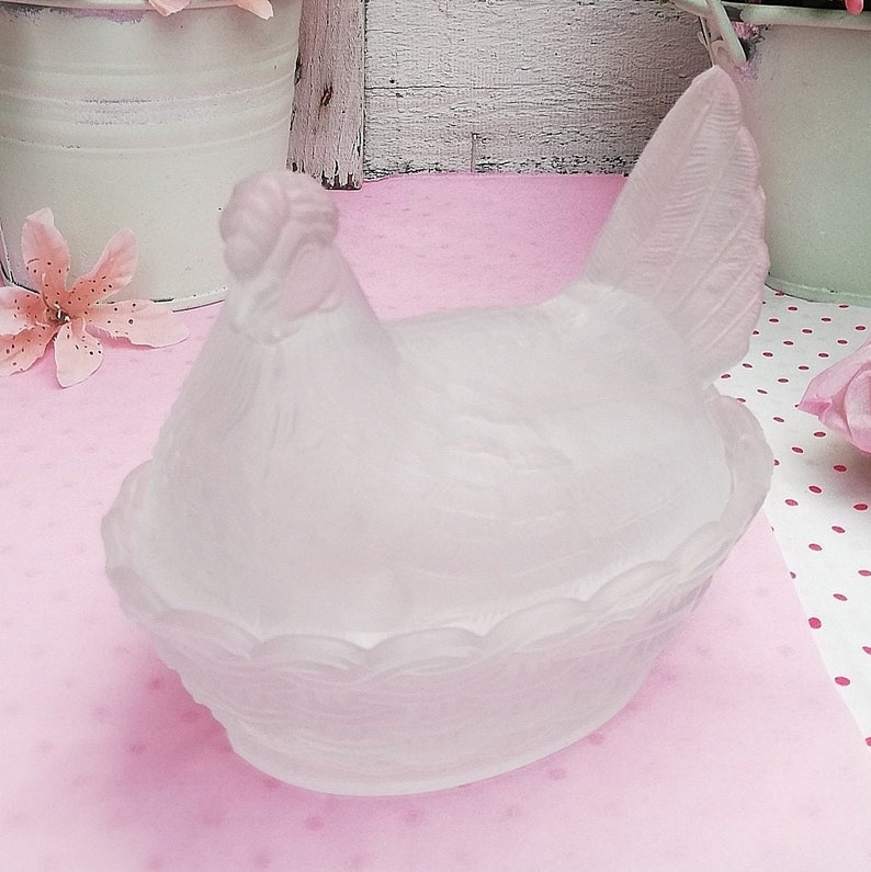 May include: A frosted glass chicken shaped dish with a pink hue. The chicken is sitting in a nest with a detailed design.