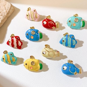 Può includere: Una collezione di ciondoli a forma di pesce colorati e smaltati in varie tonalità di blu, rosso, giallo e rosa. Ogni ciondolo presenta un anello dorato per l'attacco e un design dettagliato, tra cui pinne e occhi. I ciondoli sono disposti su una superficie bianca.