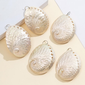 Pode incluir: Cinco pingentes de concha de abalone iridescente com argolas prateadas para fazer joias. As conchas t&ecirc;m um brilho perolado com um padr&atilde;o em espiral e uma fileira de pequenos pontos castanhos ao longo da borda. As conchas t&ecirc;m formato oval.