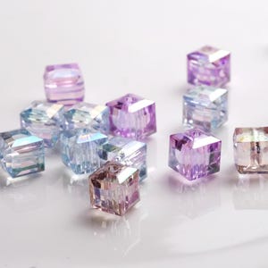 May include: A collection of iridescent glass cube beads in shades of purple and blue. The beads are faceted and have a shimmering effect.