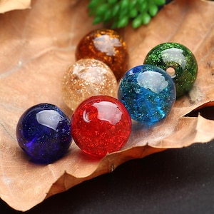 May include: A collection of six colorful glass spheres in shades of blue, red, green, gold, and amber. The spheres are arranged on a brown leaf, with a few green leaves in the background. The spheres have a speckled, glittery appearance.