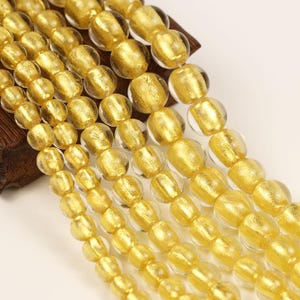 May include: Close-up of several strands of golden beads. The beads have a clear, rounded shape with a gold foil-like interior. The beads are strung together, creating a shimmering effect. The background is a light, neutral color.