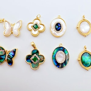 May include: A collection of gold-toned pendants featuring various designs. Included are butterfly, clover, and oval shapes, some with iridescent abalone shells and others with white or green accents. Each pendant has a loop for attaching to a chain.