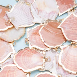 Peut inclure: Gros plan de plusieurs pendentifs en forme de coquillages avec des bords dorés. Les coquillages sont dans des tons de rose et de blanc, avec un aspect naturel et texturé. Chaque pendentif a une petite boucle pour l'attacher à une chaîne ou à un autre composant de bijou.