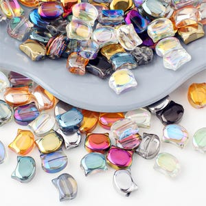 Könnte beinhalten: Eine Sammlung von irisierenden katzenförmigen Perlen in verschiedenen Farben, darunter Gold, Blau, Rosa und Klar. Die Perlen sind auf einer grauen Oberfläche verstreut und bilden eine ansprechende Darstellung für Bastelarbeiten oder Schmuckherstellung.