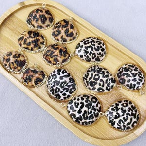 May include: Various leopard print shell pendants with gold-coloured edging, arranged on a wooden tray. The pendants are in different shapes, including scallop, round, and oval. The leopard print has black spots on a tan backdrop.