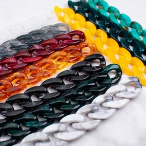 Puede incluir: Una colección de eslabones de cadena acrílicos coloridos en varios tonos, incluyendo blanco, gris, rojo, naranja, amarillo, turquesa, negro y patrones veteados. Las cadenas están dispuestas en filas paralelas, mostrando su acabado brillante y diseño grueso.