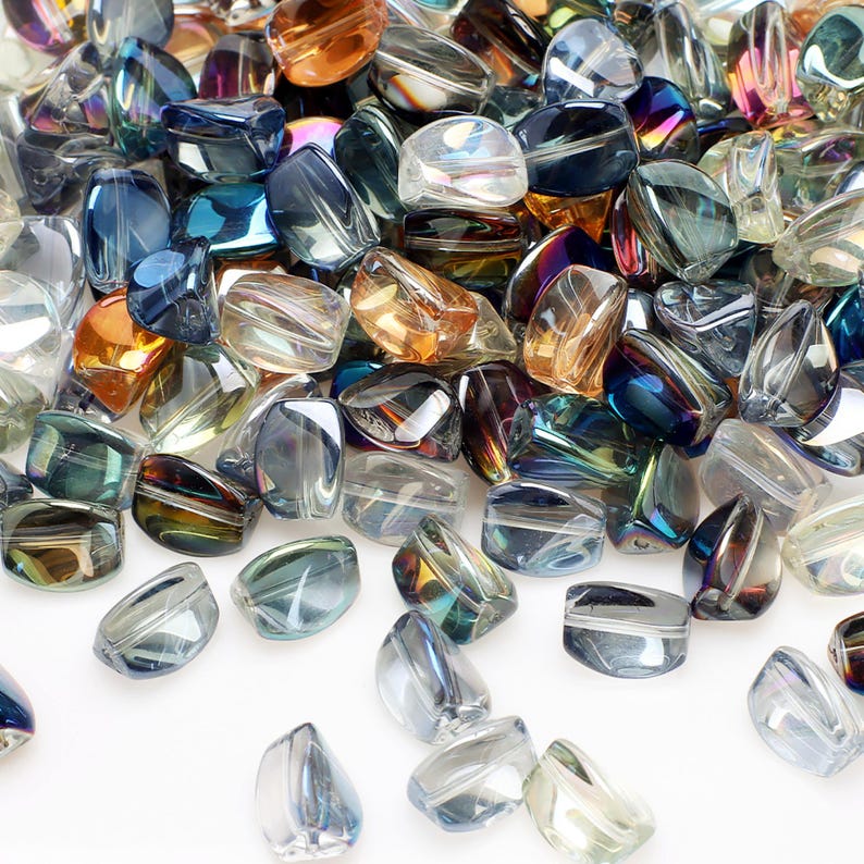 Pode incluir: Uma cole&ccedil;&atilde;o de contas de vidro iridescentes em v&aacute;rias formas e tamanhos. As contas exibem uma gama de cores, incluindo azul, &acirc;mbar e transparente, com uma qualidade brilhante e reflexiva. As contas est&atilde;o espalhadas, criando uma exibi&ccedil;&atilde;o texturizada e visualmente atraente.