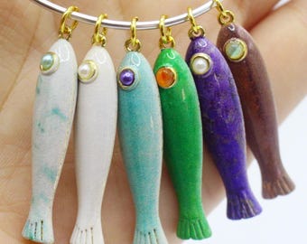 Puede incluir: Una pulsera plateada con seis colgantes en forma de pez de varios colores. Los colgantes son blancos, turquesa, verde, morado y marrón, cada uno con un ojo dorado y una perla o gema. Los peces tienen superficies texturizadas y aletas detalladas.