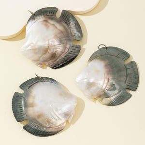 May include: Three fish-shaped pendants carved from iridescent mother-of-pearl shell. Each pendant features a smooth, pearlescent body with dark, grey-toned edges and fins, and includes a small metal jump ring attached to the top for hanging.
