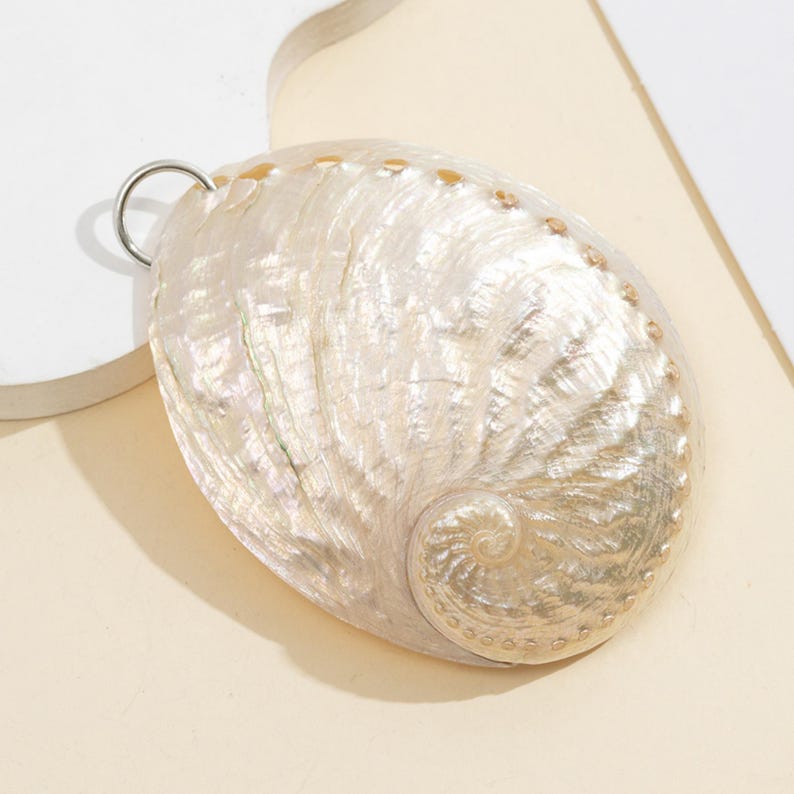 Pode incluir: Um pingente ovalado perolado com uma argola prateada. A concha iridescente apresenta um desenho em espiral e pequenos pontos castanhos na borda. A superf&iacute;cie da concha tem uma textura natural. Forma natural e org&acirc;nica.