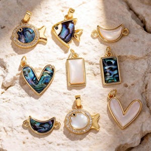 May include: A selection of gold-plated pendants in various shapes, including fish and hearts. Some pendants feature abalone shell inlays, while others have white mother-of-pearl. Several are adorned with clear crystals, offering a touch of sparkle.