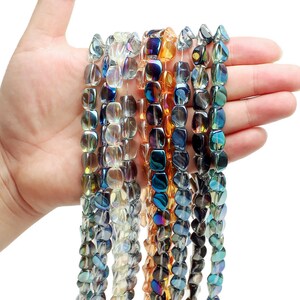 Pode incluir: Fios de contas de vidro iridescente em v&aacute;rias cores, incluindo azul, dourado e transparente. As contas, seguradas numa m&atilde;o, mostram uma forma retangular e superf&iacute;cies refletoras. Ideais para a cria&ccedil;&atilde;o de joias ou projetos de artesanato.