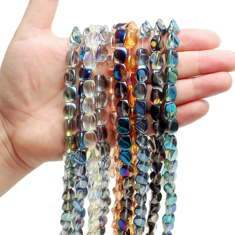 Pode incluir: Fios de contas de vidro iridescente em v&aacute;rias cores, incluindo azul, dourado e transparente. As contas, seguradas numa m&atilde;o, mostram uma forma retangular e superf&iacute;cies refletoras. Ideais para a cria&ccedil;&atilde;o de joias ou projetos de artesanato.