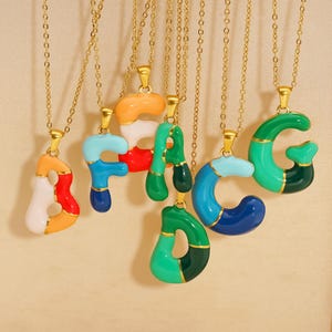 Op de afbeelding: Een verzameling goudkleurige kettingen, elk met een kleurrijke, met emaille gevulde letterhanger. De letters zijn in verschillende kleuren, waaronder rood, oranje, blauw, groen en wit, met gouden accenten. De kettingen worden tegen een neutrale achtergrond getoond.