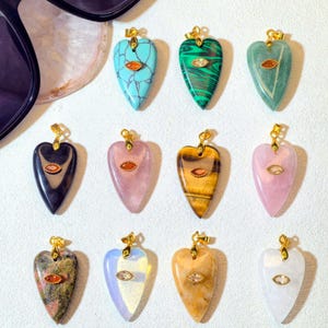 May include: A selection of heart-shaped gemstone pendants in diverse colours, such as turquoise, green, pink, and brown. Each pendant features a gold-coloured bail and a small, oval accent stone.