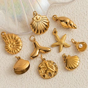 May include: A collection of gold-colored seashell charms, including a starfish, clam, and spiral shell. The charms are designed with intricate details and are displayed on a textured surface. These pendants are ideal for jewelry making.