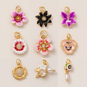 Könnte beinhalten: Neun goldfarbene Blumenanhänger mit verschiedenen Designs. Die Anhänger haben farbenfrohe Emaille-Blütenblätter in Rosa-, Lila-, Weiß- und Schwarztönen mit funkelnden Kristall- oder Perlenakzenten. Jeder Anhänger hat eine Schlaufe zum Befestigen an einer Halskette oder einem Armband.