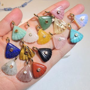 May include: A selection of gemstone pendants in diverse colours, such as blue, green, yellow, pink, and black. Each pendant features a gold-coloured bail and a small, round gemstone or crystal accent. The pendants are held in a hand.