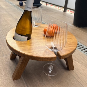May include: A circular wooden wine table with a bottle of white wine and two wine glasses. The table features a bottle holder and a glass slot. A small orange fruit rests on the table. The table is supported by three angled legs. The table is approximately 40 cm in diameter.