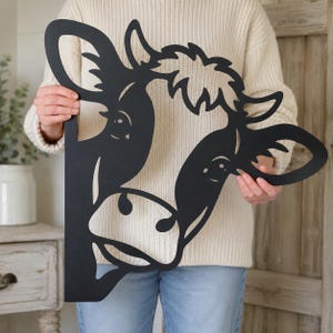 Op de afbeelding: Een plat, zwart metalen wanddecoratie in de vorm van een koeienkop. Het ontwerp toont een gestileerd silhouet van een koeiengezicht met uitgesneden details voor de ogen, oren en snuit, wat een opvallende omtrek van het dier creëert.